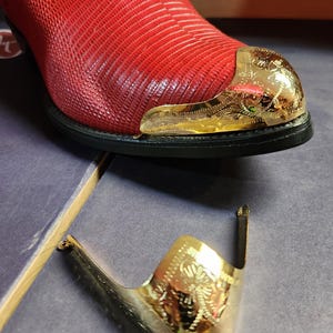 May include: A red boot with a textured lizard-skin pattern and a polished gold-tone metal toe cap featuring intricate engraved details. A matching gold-tone metal toe cap is positioned separately in the foreground on a flat surface.