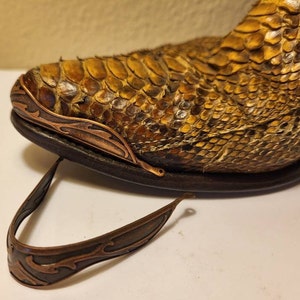 May include: Close-up of a brown and yellow snakeskin boot with a decorative copper-colored toe cap and heel guard. The metal pieces have an ornate, flowing design. The boot is on a dark sole.