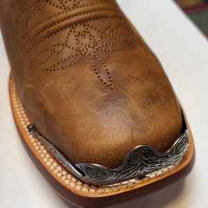 Pu&ograve; includere: Primo piano della punta di uno stivale da cowboy in pelle marrone con una punta decorativa in metallo argentato.