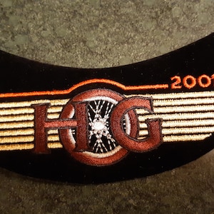 May include: A black embroidered patch with gold and red stitching. The patch features a stylized "H.O.G." logo with a motorcycle wheel in the center and the text "2001" above the logo.