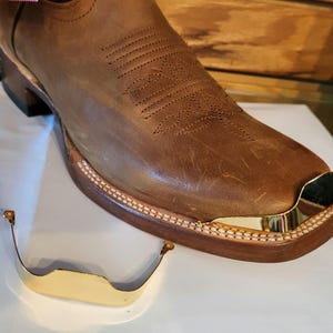 May include: Close-up of a brown leather cowboy boot with a gold metal toe cap. A separate gold metal toe cap is shown in the foreground.