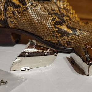 May include: A close-up of a snakeskin cowboy boot with silver metal toe and heel caps. A small bag of screws is next to the caps. The boot has a brown heel.
