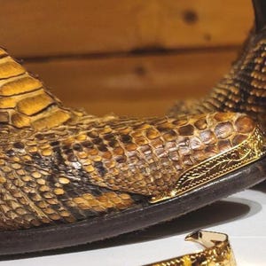 May include: A brown and black snakeskin textured boot featuring a pointed toe with an ornate gold-tone metal tip. A matching gold-tone metal toe cap with intricate engraved floral detailing is positioned in the foreground, separate from the boot.