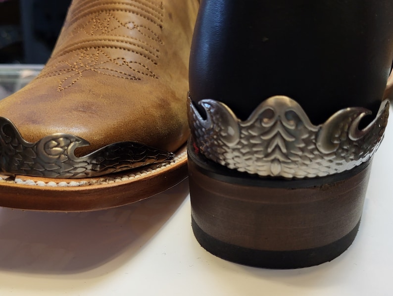 Pu&ograve; includere: Primo piano di un paio di stivali da cowboy in pelle marrone con puntali in metallo argentato. Gli stivali hanno disegni intricati sulle punte e sono usurati.
