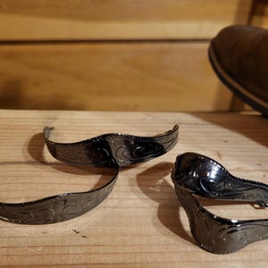 May include: Three black metal spur straps with an engraved floral design are displayed on a wooden surface. A brown leather boot is visible in the background, suggesting a Western or equestrian theme.