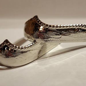 May include: Two silver spurs with intricate floral and scroll designs. The spurs have a curved shape with a small loop for attaching to a boot and a rowel. The spurs are made of a shiny, reflective metal.