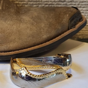 May include: Silver and gold-toned spurs with an engraved floral design. The spurs are positioned in front of a brown leather cowboy boot, showcasing a Western theme. The spurs have a decorative edge.