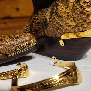 Może przedstawiać: Zbliżenie butów kowbojskich ze wzorem skóry węża w odcieniach brązu i beżu. Buty mają ciemnobrązowe obcasy i złote metalowe akcenty. Widoczne są również dwie złote ostrogi.