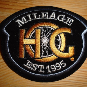 May include: Black and gold embroidered patch with the text "Mileage" above the letters "HC" and the text "Est 1995" below. The letters "HC" are surrounded by a stylized wheel.