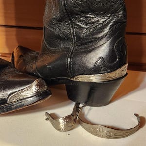 Puede incluir: Un par de botas vaqueras de cuero negro con costuras intrincadas en la caña y punteras y taloneras de metal plateado. Dos piezas decorativas de metal plateado sueltas con grabados de volutas se encuentran en el primer plano.