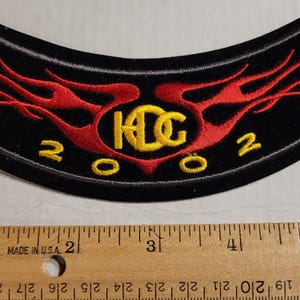 May include: Black, curved patch with red flame design and yellow text. The text includes "HCC" in a stylized font and the year "2002". The patch is approximately 5 inches wide.
