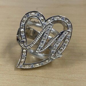 Peut inclure: Une bague en argent avec un design en forme de cœur représentant la lettre "M" entourée de petites pierres claires.