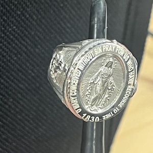 May include: A silver ring with a detailed image of the Virgin Mary. The ring has the inscription "O Mary Conceived Without Sin Pray For Us Who Have Recourse To Thee 1830".