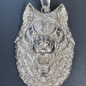 May include: A silver-toned pendant featuring a highly detailed, three-dimensional wolf head with an open mouth, bared teeth, and textured fur. The pendant includes a polished bail at the top for attachment to a chain, showcasing a metallic finish.