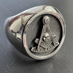 May include: A silver-toned ring with a prominent oval face. The face features a detailed emblem with a skull, compass, and square design. The ring has a polished, reflective surface.