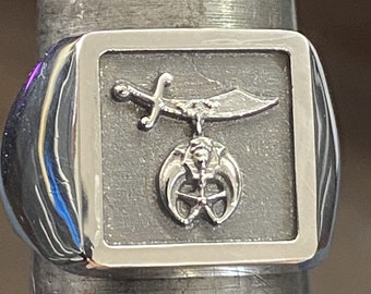 Può includere: Un anello d'argento con un designo quadrato che presenta una spada e una mezzaluna con una stella al centro.