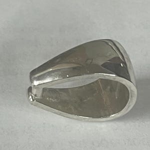 Pode incluir: Biela de metal prateada para pendente com uma superfície lisa e arredondada. A argola é projetada para prender um pendente a uma corrente ou cordão.