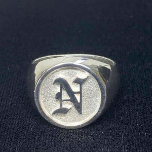 May include: A silver ring with a raised, circular face. The face features a textured background and a gothic-style letter "N" in the center. The ring has a polished, reflective finish.
