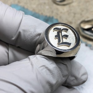 May include: A silver signet ring with a circular face. The face features a raised, stylized letter "E" in a dark color against a lighter background. The ring is held by a gloved hand, with other rings visible in the background.