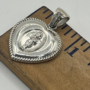 May include: Silver heart-shaped pendant with a detailed angel design and a row of clear cubic zirconia stones on the bail.