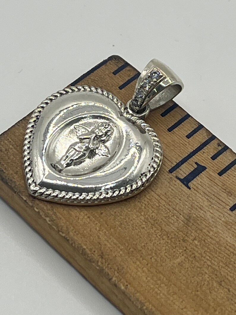 May include: Silver heart-shaped pendant with a detailed angel design and a row of clear cubic zirconia stones on the bail.