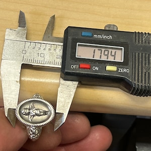 K&ouml;nnte beinhalten: Ein silberner Ring mit einem ovalen Design, das einen Cherub mit Fl&uuml;geln zeigt. Der Ring wird mit einer Schieblehre gemessen, die 17,94 mm anzeigt.