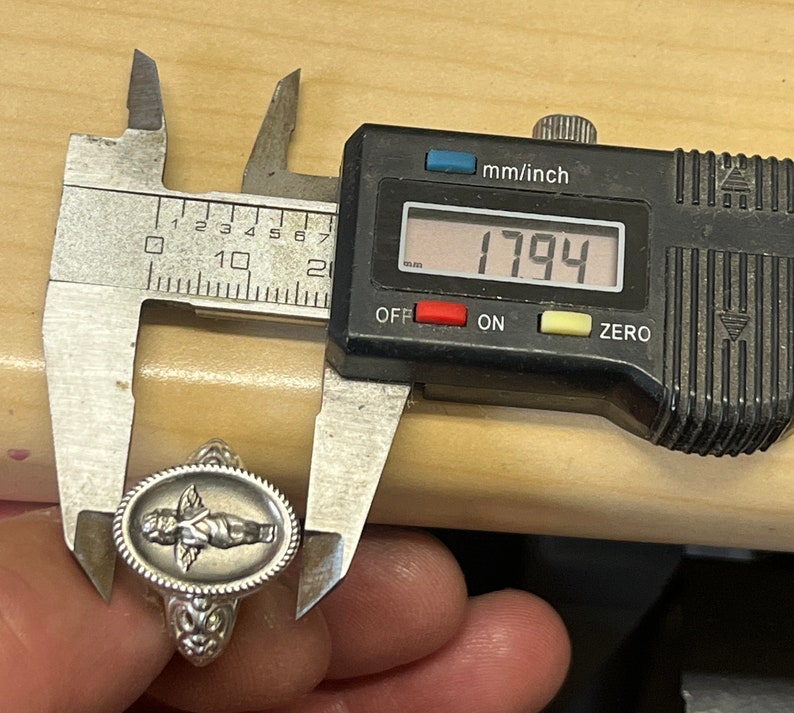 K&ouml;nnte beinhalten: Ein silberner Ring mit einem ovalen Design, das einen Cherub mit Fl&uuml;geln zeigt. Der Ring wird mit einer Schieblehre gemessen, die 17,94 mm anzeigt.