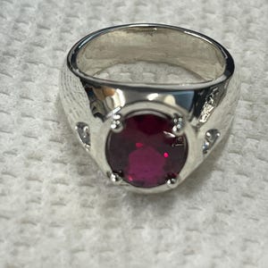 May include: A silver ring with a large, oval-shaped, dark red gemstone surrounded by smaller, clear gemstones.