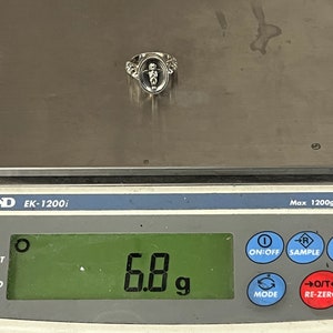 K&ouml;nnte beinhalten: Ein silberner Ring mit einem schwarzen ovalen Stein, der ein detailliertes Design eines Cherubs zeigt. Der Ring befindet sich auf einer digitalen Waage, die 6,8 Gramm anzeigt.