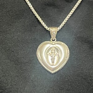 May include: A silver heart-shaped pendant necklace with a small angel design inside the heart. The chain is a silver curb chain.