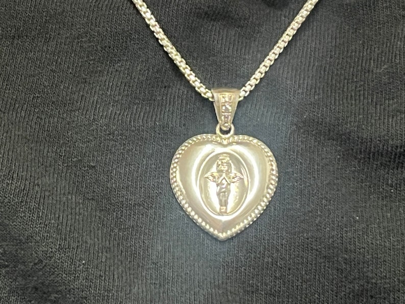 May include: A silver heart-shaped pendant necklace with a small angel design inside the heart. The chain is a silver curb chain.