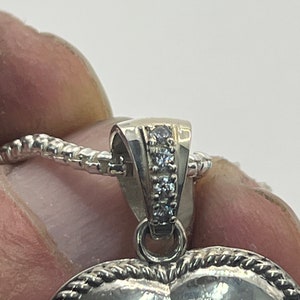 May include: Silver heart-shaped pendant with a rope-like border and a row of small clear stones on the bail.
