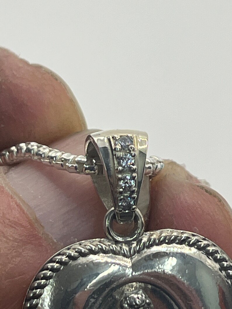 May include: Silver heart-shaped pendant with a rope-like border and a row of small clear stones on the bail.