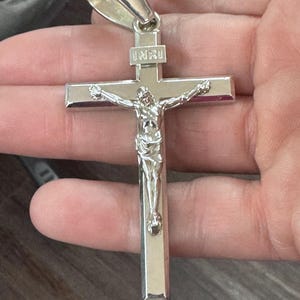 Może przedstawiać: Srebrny wisiorek z krzyżem. Krzyż przedstawia szczegółowe przedstawienie Jezusa Chrystusa i napis „INRI” na górze. Wisiorek jest trzymany przez srebrne kółko.