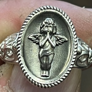 K&ouml;nnte beinhalten: Ein silberner Ring mit einem ovalen Design, das eine detaillierte Skulptur eines Engels mit Fl&uuml;geln, ineinander verschr&auml;nkten H&auml;nden zum Gebet und einem strukturierten Rand zeigt.