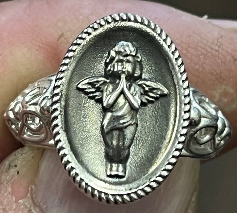 K&ouml;nnte beinhalten: Ein silberner Ring mit einem ovalen Design, das eine detaillierte Skulptur eines Engels mit Fl&uuml;geln, ineinander verschr&auml;nkten H&auml;nden zum Gebet und einem strukturierten Rand zeigt.
