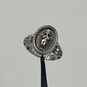 K&ouml;nnte beinhalten: Ein silberner Ring mit einem ovalen Design, das einen Cherub mit Fl&uuml;geln zeigt. Der Cherub h&auml;lt eine Blume und hat ein zartes, detailliertes Design.