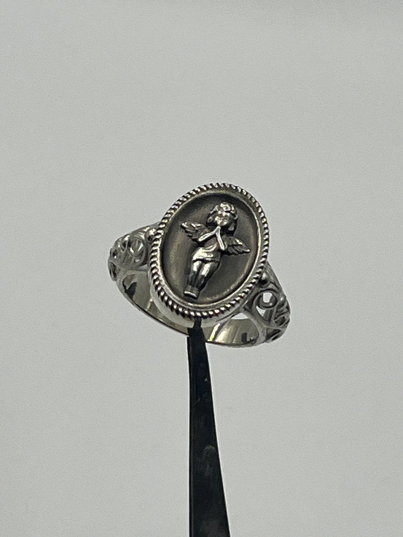 K&ouml;nnte beinhalten: Ein silberner Ring mit einem ovalen Design, das einen Cherub mit Fl&uuml;geln zeigt. Der Cherub h&auml;lt eine Blume und hat ein zartes, detailliertes Design.