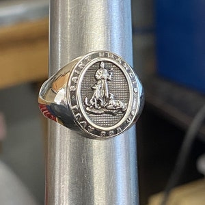 May include: A silver ring with an oval-shaped design featuring a raised image of Saint Francis of Assisi holding a bird. The ring is inscribed with "SAINT FRANCIS OF ASSISI".