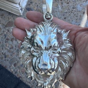 Puede incluir: Un colgante plateado con un diseño detallado de cabeza de león. El colgante tiene una argolla grande para una cadena. La cara del león está intrincadamente esculpida con una melena y rasgos faciales. El colgante se sostiene en una mano.