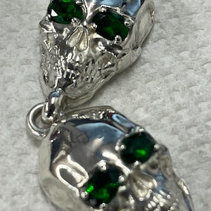 May include: Two silver skull pendants with green gemstone eyes. The skulls are detailed and have a shiny finish.