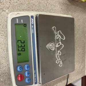May include: A silver chain necklace with a heart-shaped pendant, weighing 23.99 grams, as shown on a digital scale.