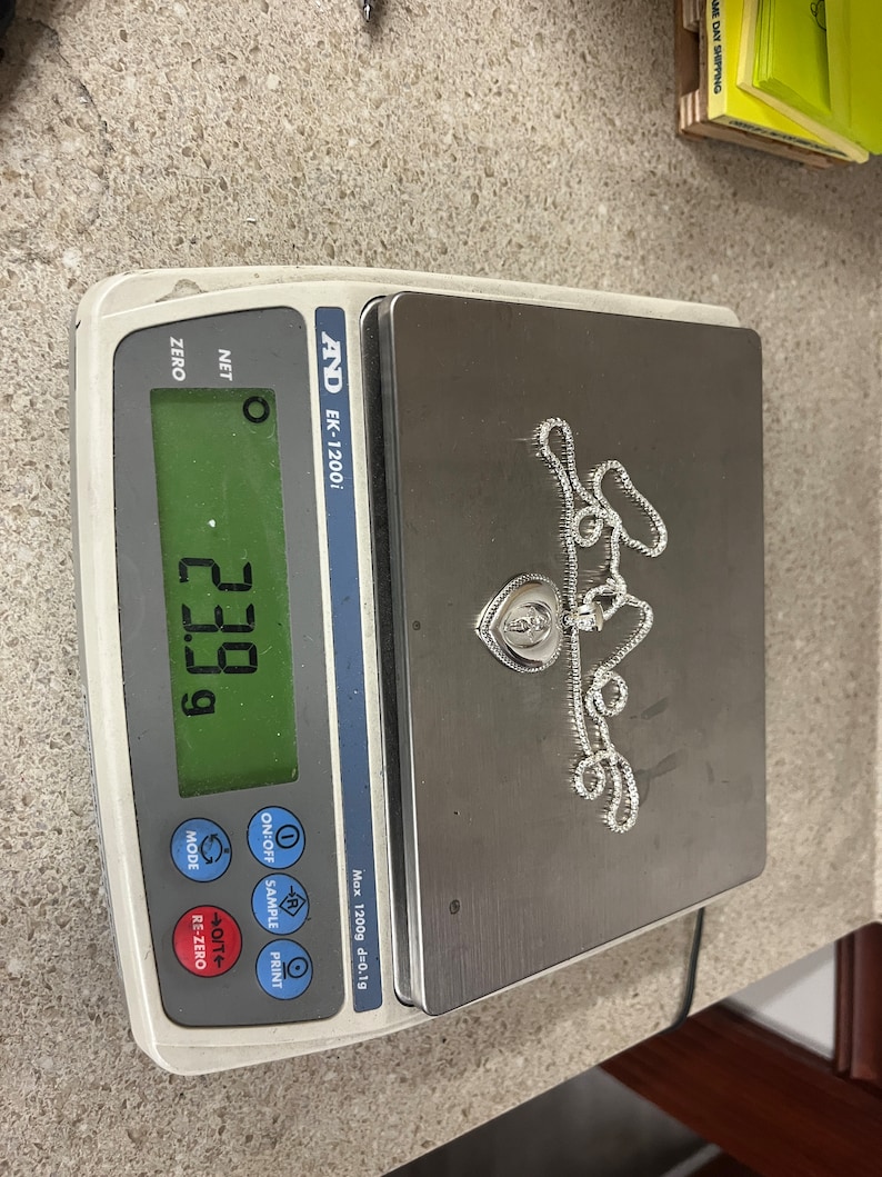 May include: A silver chain necklace with a heart-shaped pendant, weighing 23.99 grams, as shown on a digital scale.