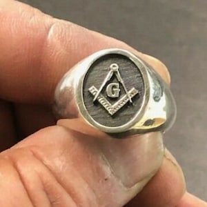 May include: A silver ring with a black and gold Masonic symbol. The symbol is a square and compass with a 'G' in the center.
