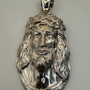 May include: Silver pendant featuring a detailed, three-dimensional depiction of Jesus Christ's face with a crown of thorns. The pendant is designed to be worn as a necklace.