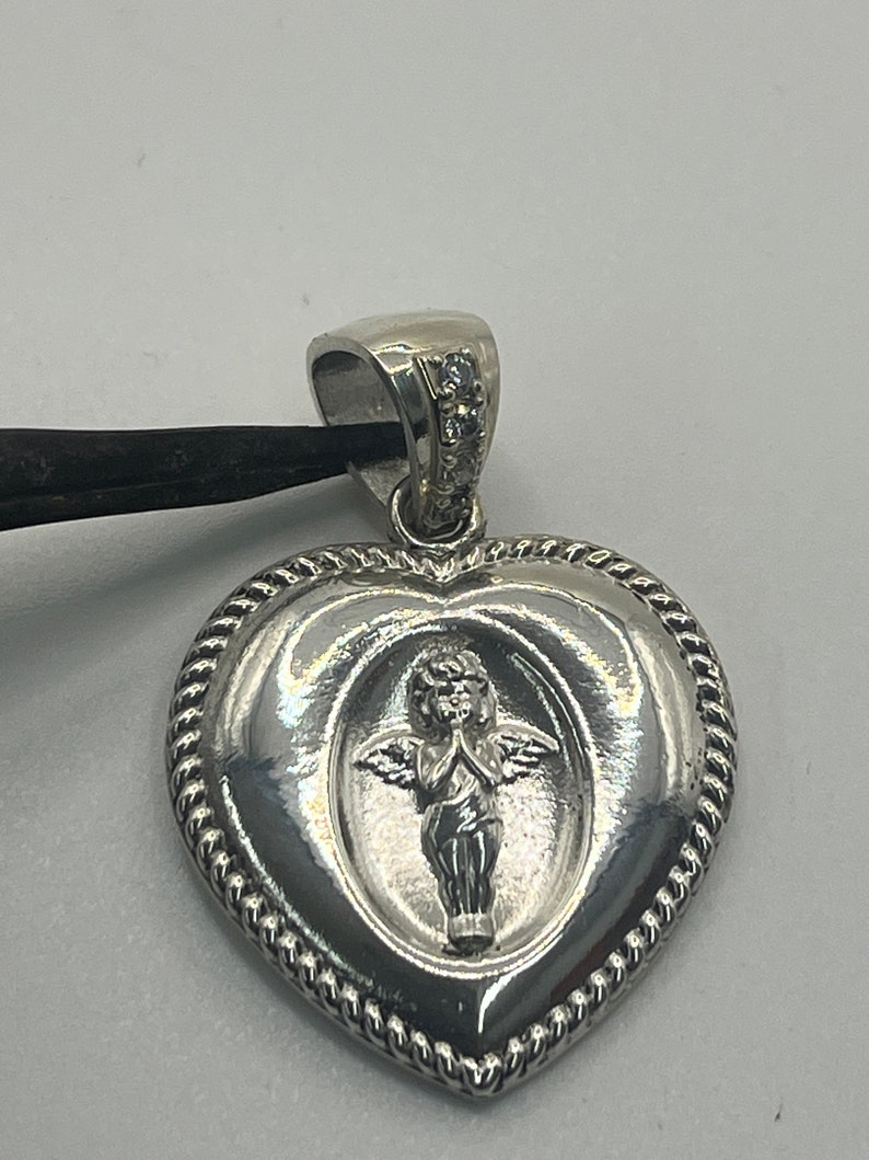 May include: Silver heart-shaped pendant with a detailed angel design and small clear stones on the bail.