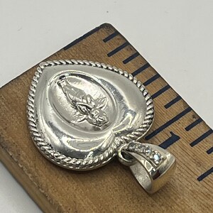 May include: Silver heart-shaped pendant with a detailed design of a figure with wings and a flower. The pendant has a rope-like border and a bail with clear stones.