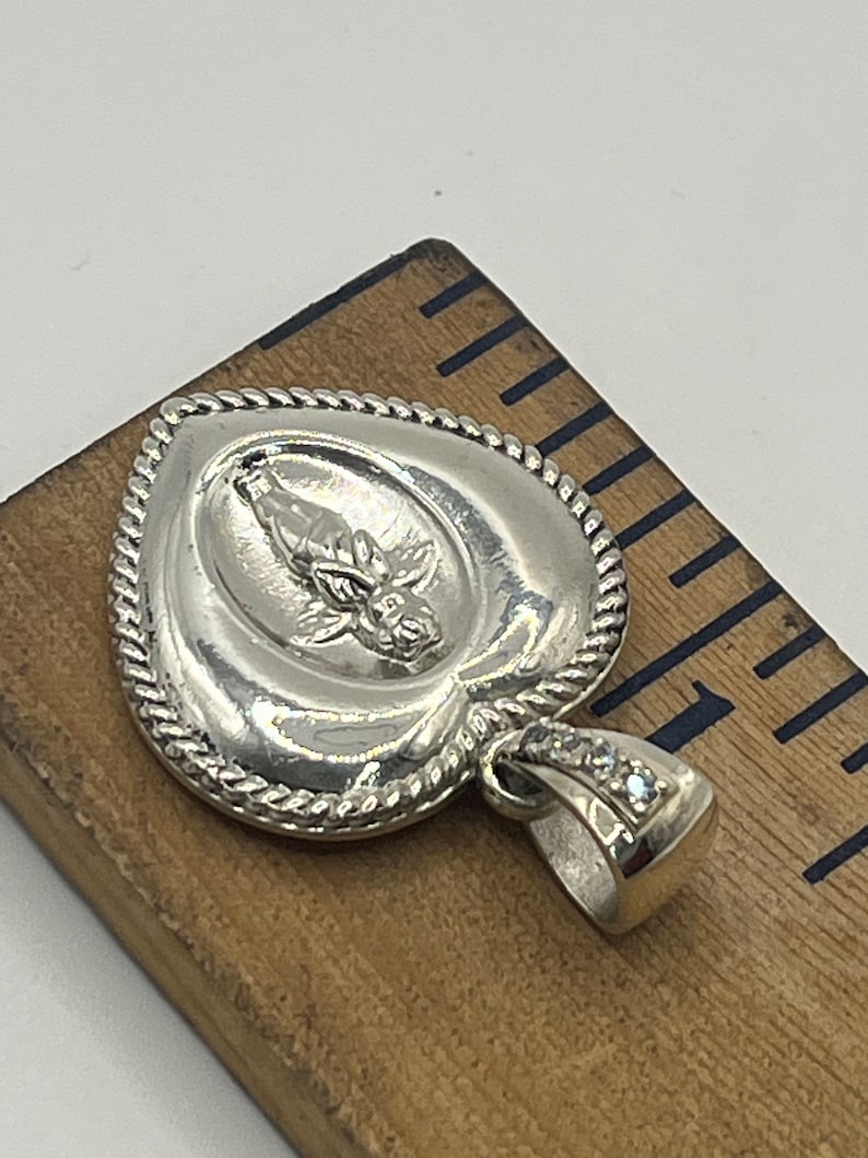 May include: Silver heart-shaped pendant with a detailed design of a figure with wings and a flower. The pendant has a rope-like border and a bail with clear stones.