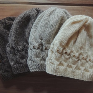May include: Four knitted beanies in shades of gray and cream are displayed. Each hat has a textured pattern around the brim and is adorned with small, decorative buttons. The hats are arranged on a wooden surface.