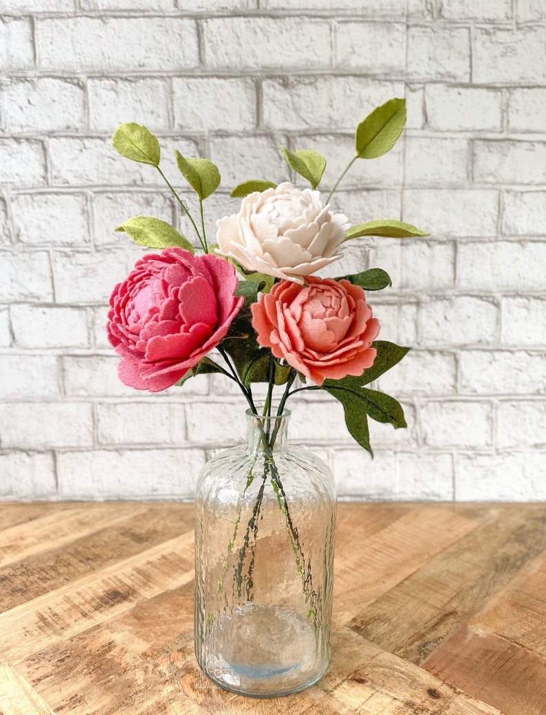 May include: A clear glass vase holds a bouquet of felt peonies. The arrangement features a vibrant mix of colors, including a deep pink, coral, and cream-colored blooms, complemented by green leaves. The vase sits on a wooden surface against a white brick wall.